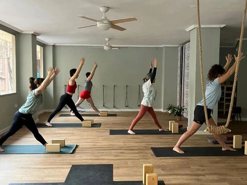 Persona realizando una postura de yoga inclinada hacia adelante con las manos juntas, transmitiendo calma y atención enfocada; aparece una etiqueta con la palabra Principiante.